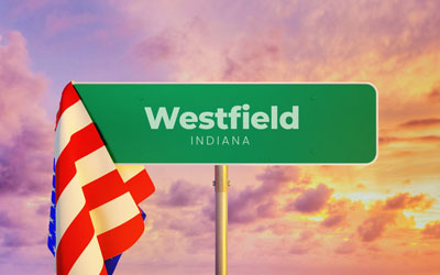 Green highway sign saying Westfield Indiana, with an AMerican flag draped on the left side against an orange sunset sky.