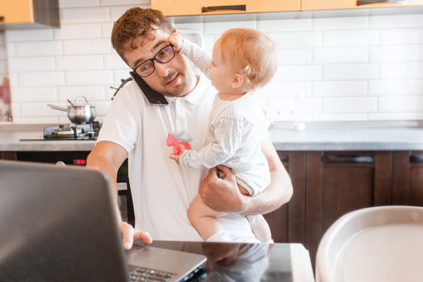 Dad working from home holding a baby who is grabbing his eyeglasses while he is standing and trying to use his laptop on the kitchen counter.