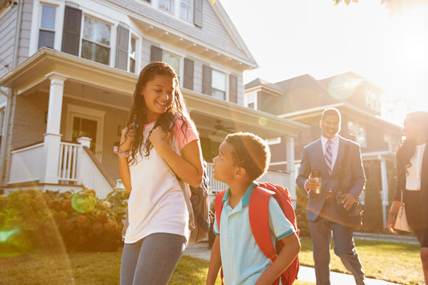 Two children with backpacks walking on the sidewalk in front of what appear to be their parents dressed for work.